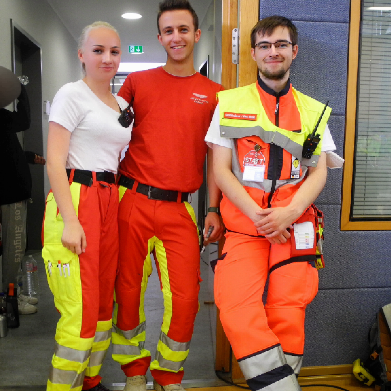 Drei Personen in Arbeitskleidung, lächelnd, stehen in einer Turnhalle als Sanitäter*innen bereit und sichern eine Sportveranstaltung ab.